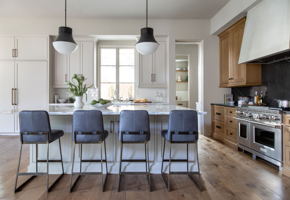 Inspiration for a large transitional l-shaped light wood floor and brown floor eat-in kitchen remodel in Houston with a farmhouse sink, recessed-panel cabinets, light wood cabinets, marble countertops, white backsplash, ceramic backsplash, paneled appliances, an island and black countertops