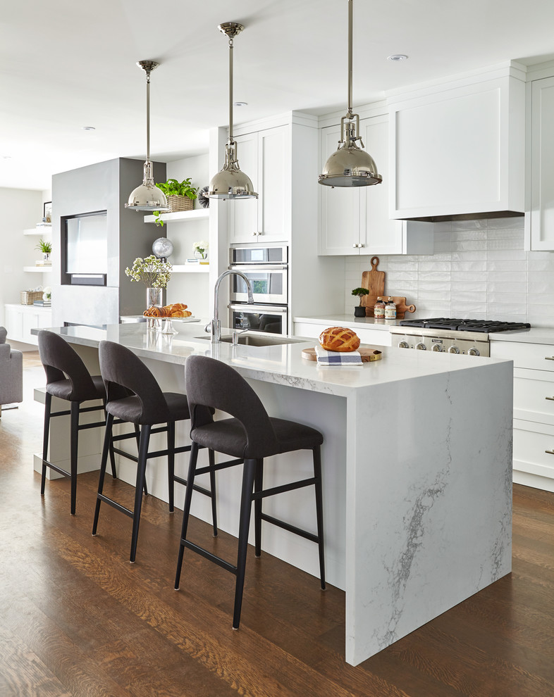 This is an example of a mid-sized transitional galley open plan kitchen in Toronto with a single-bowl sink, shaker cabinets, quartz benchtops, white splashback, porcelain splashback, stainless steel appliances, medium hardwood floors, with island, brown floor and white cabinets.
