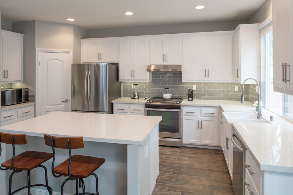 Photo of a mid-sized modern u-shaped kitchen in San Diego with a farmhouse sink, shaker cabinets, white cabinets, quartz benchtops, grey splashback, subway tile splashback, stainless steel appliances, plywood floors and with island.