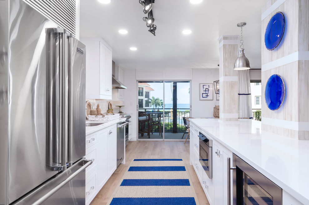 Photo of a mid-sized beach style galley open plan kitchen in Miami with an undermount sink, shaker cabinets, white cabinets, quartz benchtops, white splashback, ceramic splashback, stainless steel appliances, light hardwood floors, with island, beige floor and white benchtop.
