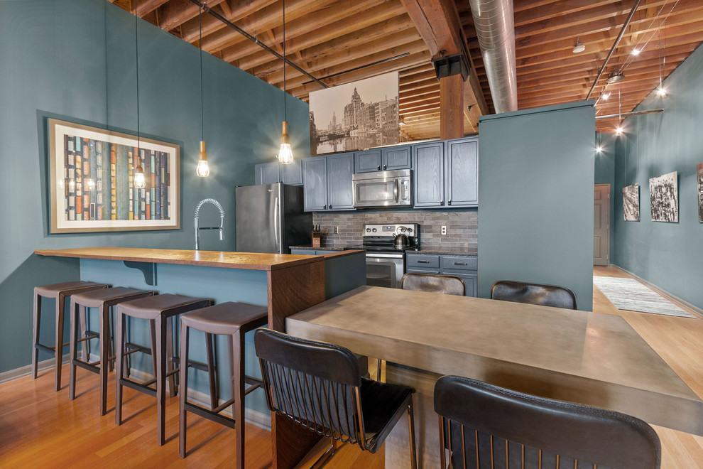 Mid-sized industrial galley open plan kitchen in Milwaukee with an undermount sink, recessed-panel cabinets, blue cabinets, granite benchtops, black splashback, ceramic splashback, stainless steel appliances, medium hardwood floors, a peninsula, brown floor and grey benchtop.