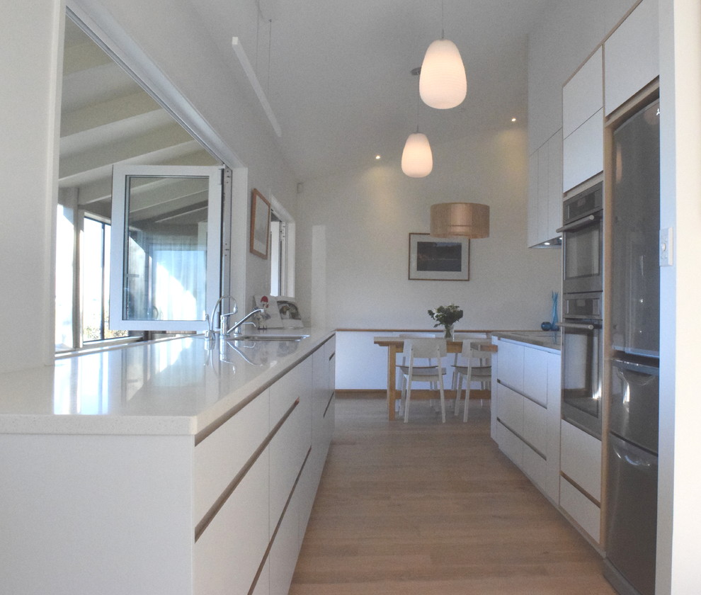 Photo of a mid-sized scandinavian galley open plan kitchen in Christchurch with an undermount sink, flat-panel cabinets, white cabinets, solid surface benchtops, multi-coloured splashback, glass sheet splashback, stainless steel appliances, medium hardwood floors, no island and brown floor.