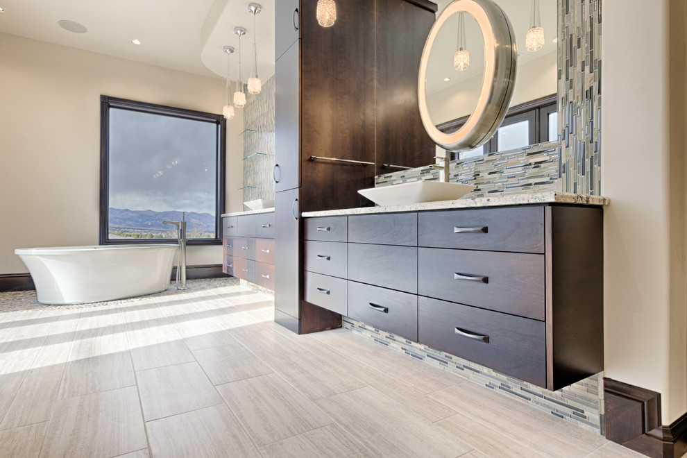 This is an example of an expansive modern master bathroom in Denver with flat-panel cabinets, dark wood cabinets, a freestanding tub, a two-piece toilet, multi-coloured tile, glass sheet wall, beige walls, porcelain floors, a vessel sink, granite benchtops, beige floor and a hinged shower door.