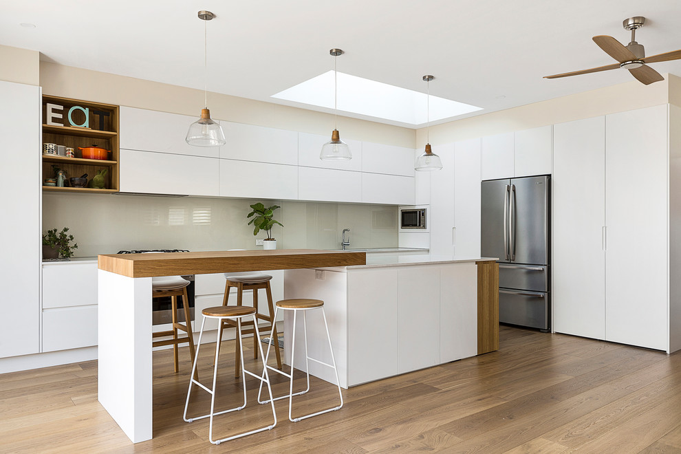 Design ideas for a large scandinavian l-shaped open plan kitchen in Sydney with an undermount sink, flat-panel cabinets, white cabinets, quartz benchtops, white splashback, glass sheet splashback, stainless steel appliances, medium hardwood floors, with island, white benchtop and brown floor.