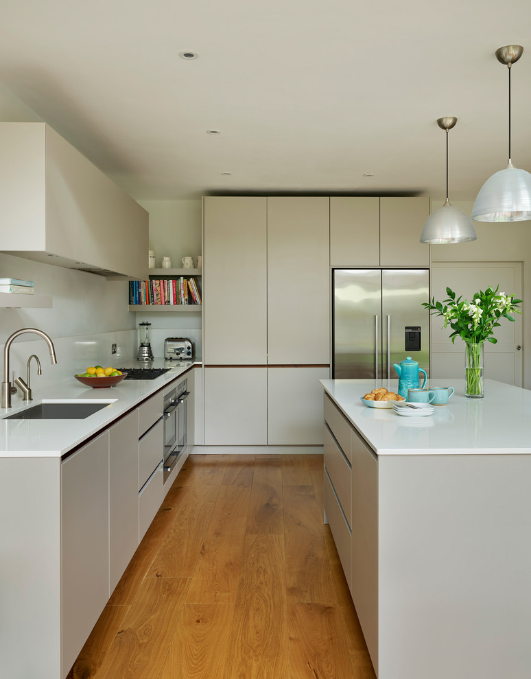 Design ideas for a large contemporary l-shaped kitchen in London with flat-panel cabinets, solid surface benchtops, medium hardwood floors, with island, white benchtop, white splashback, stainless steel appliances, an undermount sink and grey cabinets.
