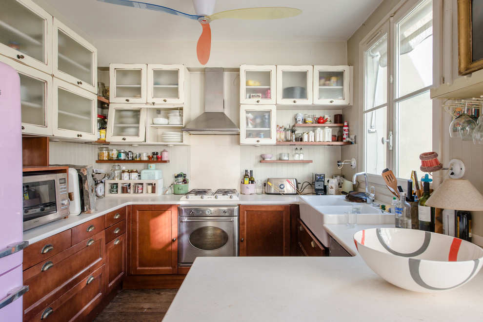 This is an example of an eclectic u-shaped kitchen in Montpellier with a peninsula, a farmhouse sink, glass-front cabinets, stainless steel appliances, dark hardwood floors, brown floor, beige cabinets, beige splashback and beige benchtop.