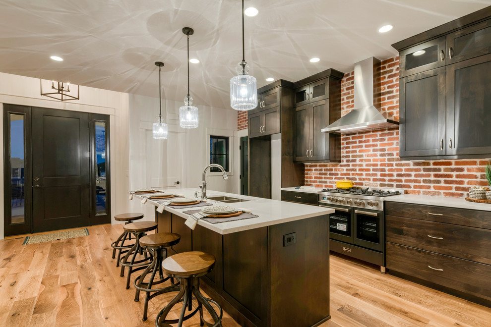 Mid-sized industrial single-wall open plan kitchen in Denver with a double-bowl sink, shaker cabinets, dark wood cabinets, quartz benchtops, brown splashback, brick splashback, stainless steel appliances, medium hardwood floors, with island, brown floor and white benchtop.