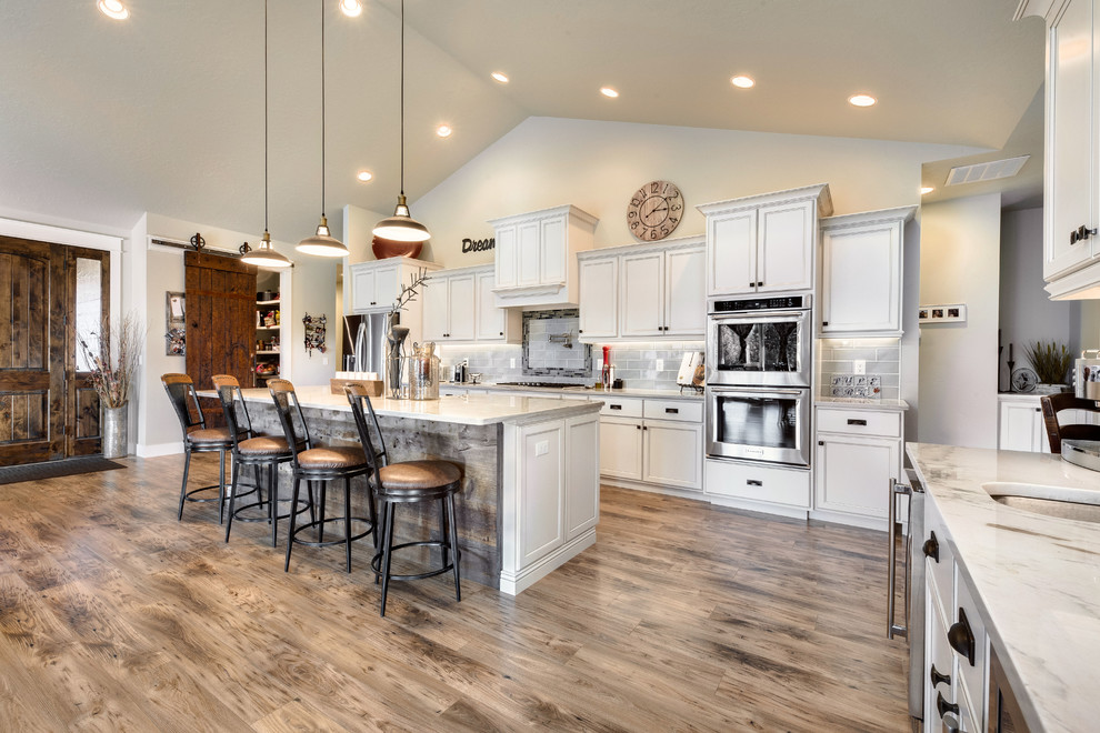 Design ideas for a large country l-shaped open plan kitchen in Orange County with an undermount sink, shaker cabinets, white cabinets, grey splashback, subway tile splashback, stainless steel appliances, medium hardwood floors, with island, brown floor and white benchtop.