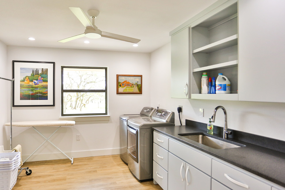 Mid-sized midcentury single-wall dedicated laundry room in Austin with an undermount sink, flat-panel cabinets, grey cabinets, solid surface benchtops, white walls, light hardwood floors, a side-by-side washer and dryer and black benchtop.