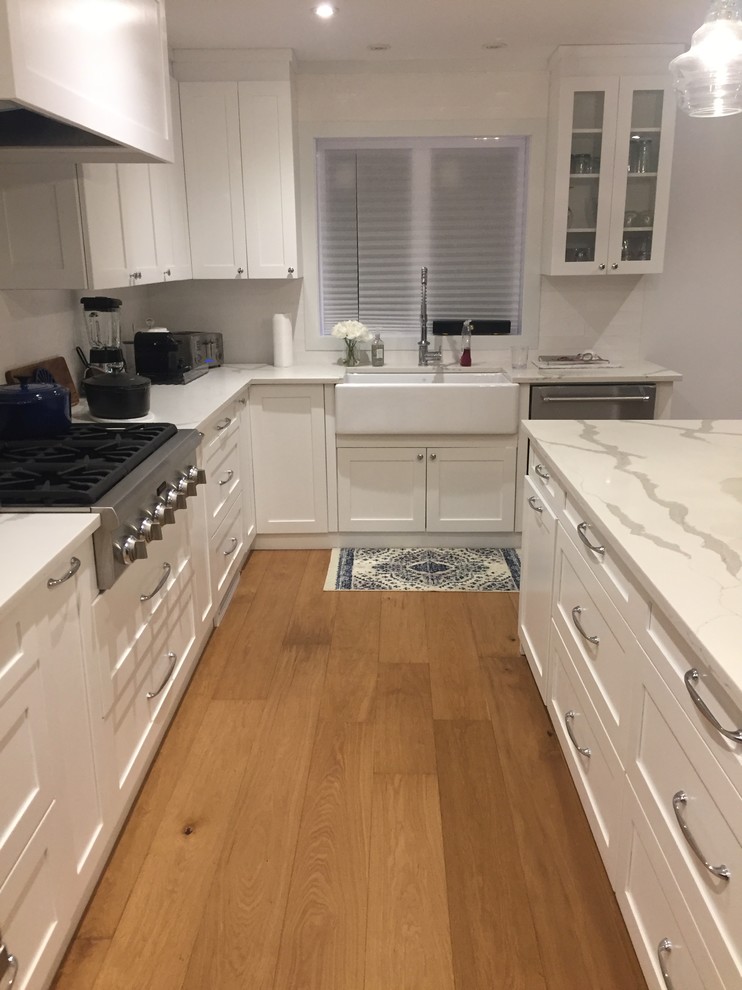 Inspiration for a large country l-shaped eat-in kitchen in Montreal with a farmhouse sink, shaker cabinets, white cabinets, quartz benchtops, white splashback, porcelain splashback, stainless steel appliances, medium hardwood floors, with island and beige floor.