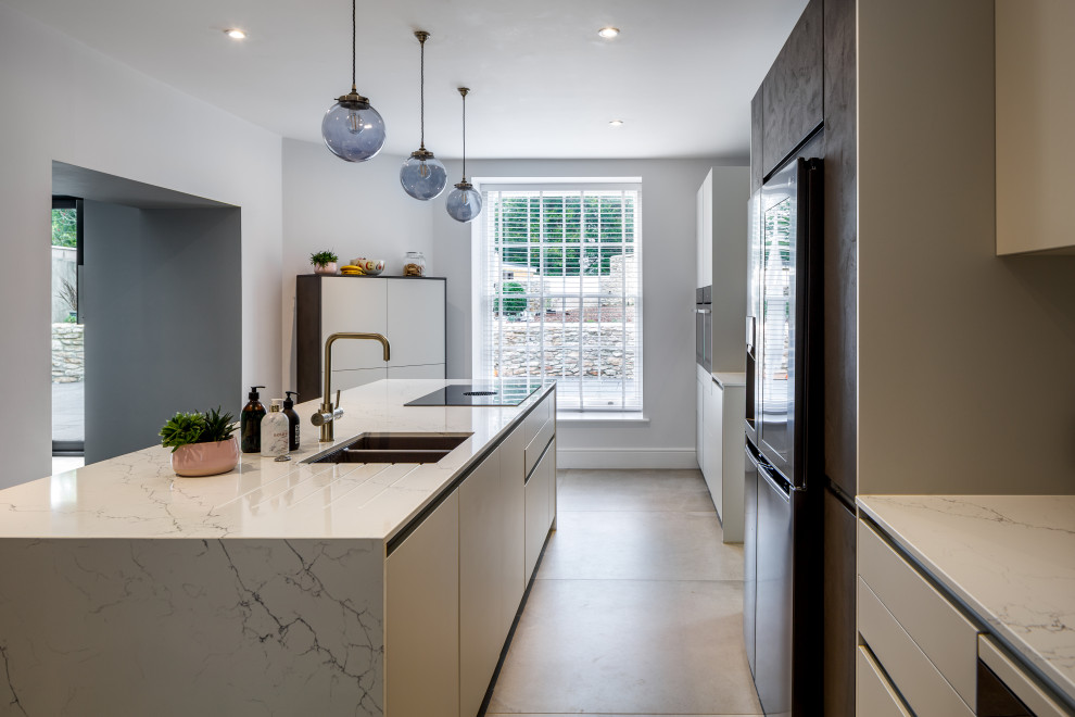 Photo of a mid-sized modern galley open plan kitchen in Devon with an integrated sink, flat-panel cabinets, white cabinets, granite benchtops, black appliances, limestone floors, with island, beige floor and white benchtop.
