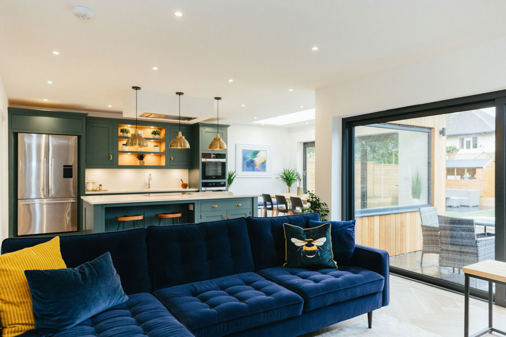 Large contemporary l-shaped open plan kitchen in London with a built-in sink, shaker cabinets, blue cabinets, white splashback, black appliances, light hardwood flooring, an island, beige floors, white worktops and feature lighting.