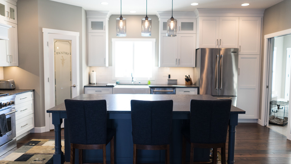Large transitional u-shaped eat-in kitchen photo with a farmhouse sink, flat-panel cabinets, white cabinets, granite countertops, white backsplash, ceramic backsplash, stainless steel appliances and an island