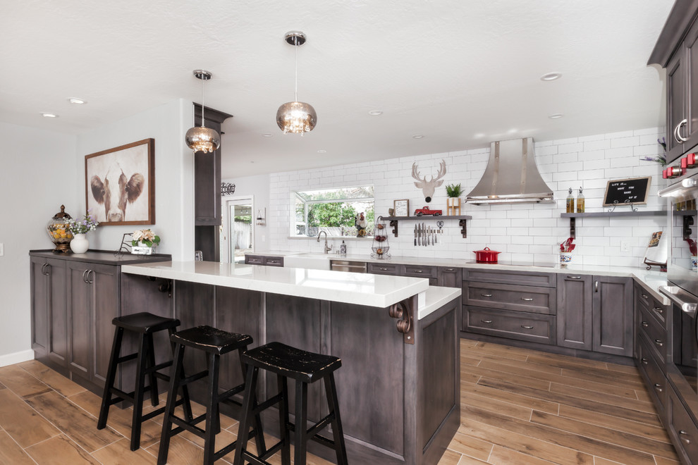 This is an example of a country kitchen in San Francisco with a farmhouse sink, subway tile splashback, stainless steel appliances, porcelain floors, shaker cabinets, brown cabinets, white splashback, a peninsula, brown floor and white benchtop.