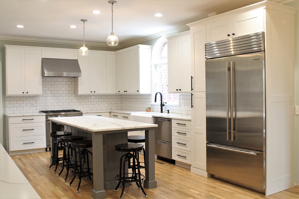 Inspiration for a large transitional galley light wood floor eat-in kitchen remodel with a farmhouse sink, shaker cabinets, white cabinets, quartz countertops, white backsplash, ceramic backsplash, stainless steel appliances, an island and white countertops
