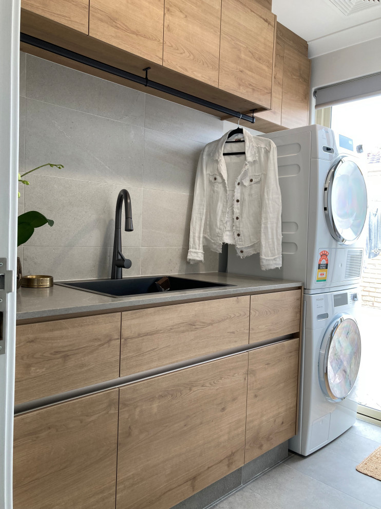 Design ideas for a mid-sized beach style galley dedicated laundry room in Perth with a drop-in sink, flat-panel cabinets, medium wood cabinets, quartz benchtops, grey splashback, porcelain splashback, grey walls, porcelain floors, a stacked washer and dryer and grey benchtop.
