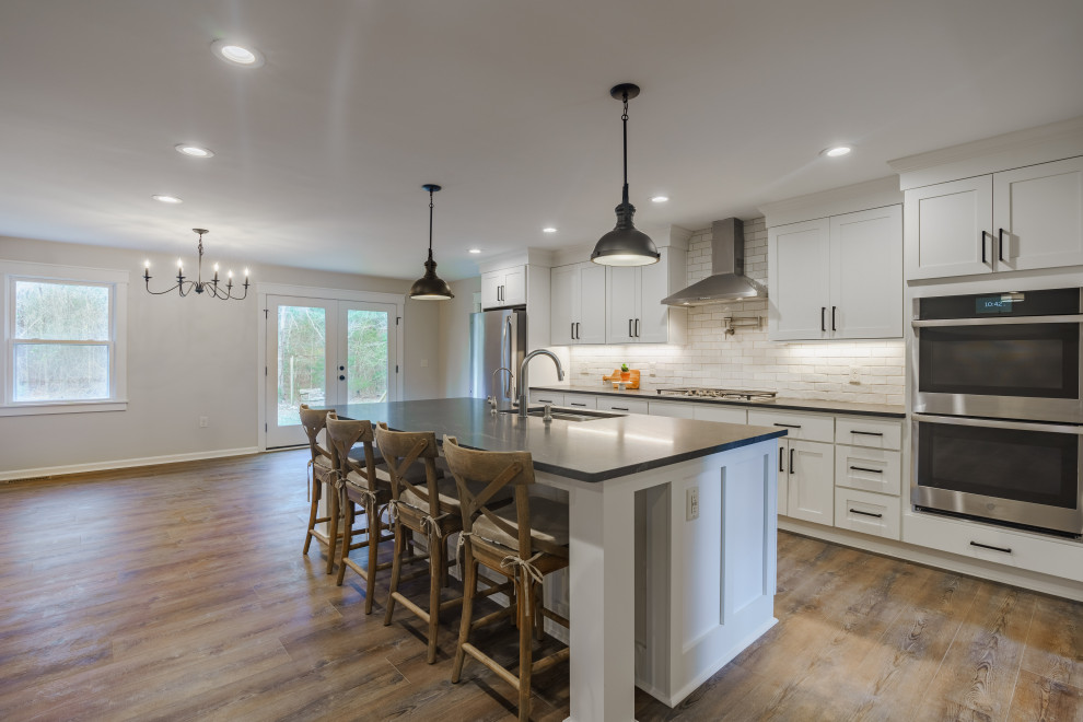 Inspiration for a mid-sized country l-shaped eat-in kitchen in Other with a drop-in sink, shaker cabinets, white cabinets, white splashback, ceramic splashback, stainless steel appliances, medium hardwood floors, with island, brown floor and vaulted.