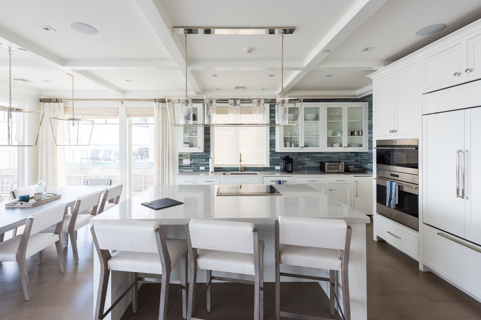 This is an example of a mid-sized beach style l-shaped open plan kitchen in New York with an undermount sink, glass-front cabinets, white cabinets, quartz benchtops, blue splashback, glass tile splashback, stainless steel appliances, medium hardwood floors, with island, brown floor and grey benchtop.