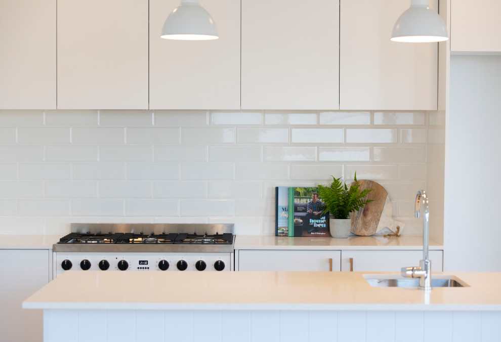 Photo of a country l-shaped open plan kitchen in Wollongong with a drop-in sink, white cabinets, quartz benchtops, white splashback, subway tile splashback, white appliances, light hardwood floors, with island and white benchtop.