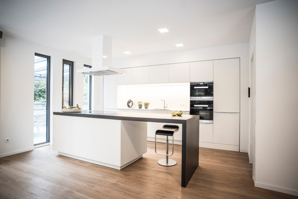 Photo of an expansive contemporary open plan kitchen in Other with flat-panel cabinets, white cabinets, white splashback, black appliances, an island and brown floors.
