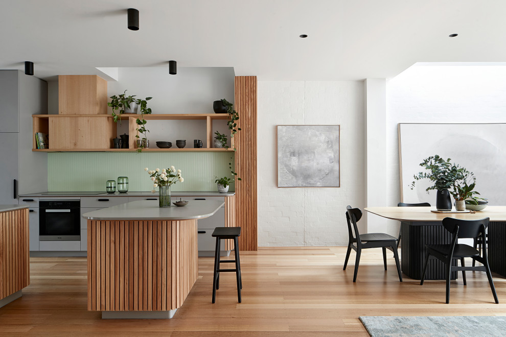 Mid-sized contemporary u-shaped open plan kitchen in Melbourne with flat-panel cabinets, grey cabinets, quartz benchtops, green splashback, glass sheet splashback, black appliances, light hardwood floors, with island, brown floor and grey benchtop.