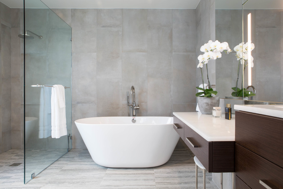 This is an example of a large contemporary master bathroom in Los Angeles with flat-panel cabinets, brown cabinets, a freestanding tub, an open shower, a wall-mount toilet, gray tile, porcelain tile, grey walls, porcelain floors, an undermount sink, glass benchtops, grey floor, an open shower and white benchtops.