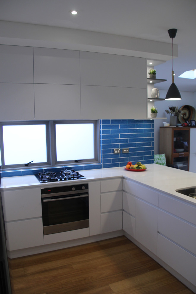 Inspiration for a mid-sized contemporary u-shaped kitchen pantry in Sydney with an undermount sink, recessed-panel cabinets, white cabinets, solid surface benchtops, blue splashback, ceramic splashback, stainless steel appliances, medium hardwood floors, with island, brown floor and white benchtop.