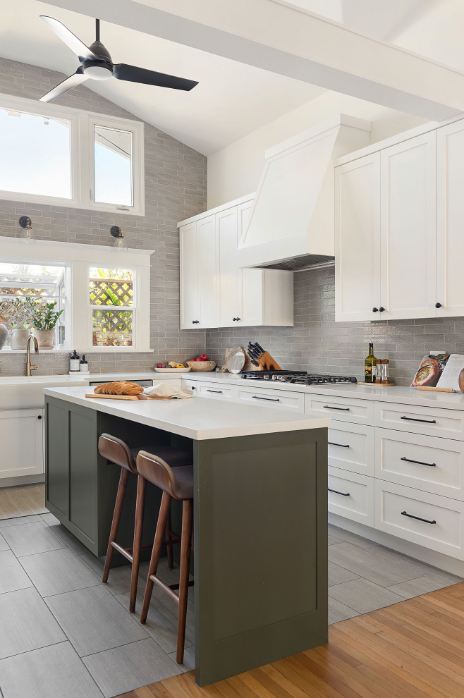 Mid-sized arts and crafts u-shaped open plan kitchen in San Diego with a farmhouse sink, shaker cabinets, white cabinets, quartz benchtops, grey splashback, ceramic splashback, stainless steel appliances, porcelain floors, with island, grey floor and white benchtop.