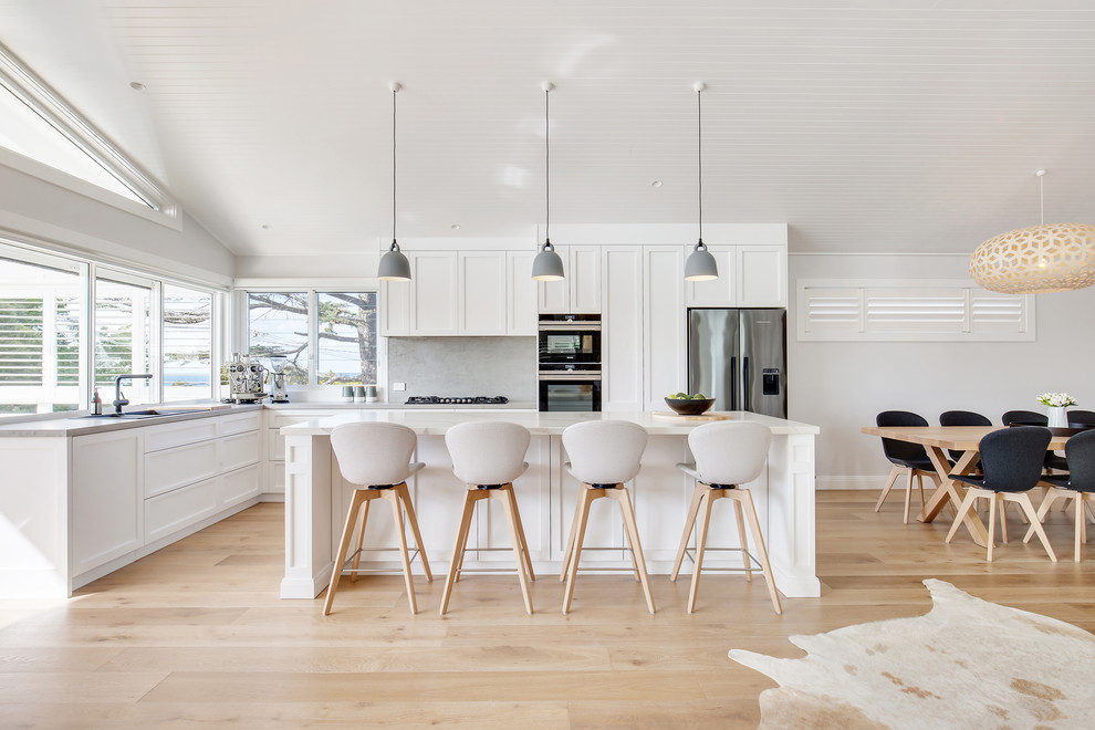 Photo of a mid-sized beach style l-shaped open plan kitchen in Sydney with a double-bowl sink, shaker cabinets, white cabinets, quartz benchtops, grey splashback, stainless steel appliances, medium hardwood floors, with island, brown floor and white benchtop.