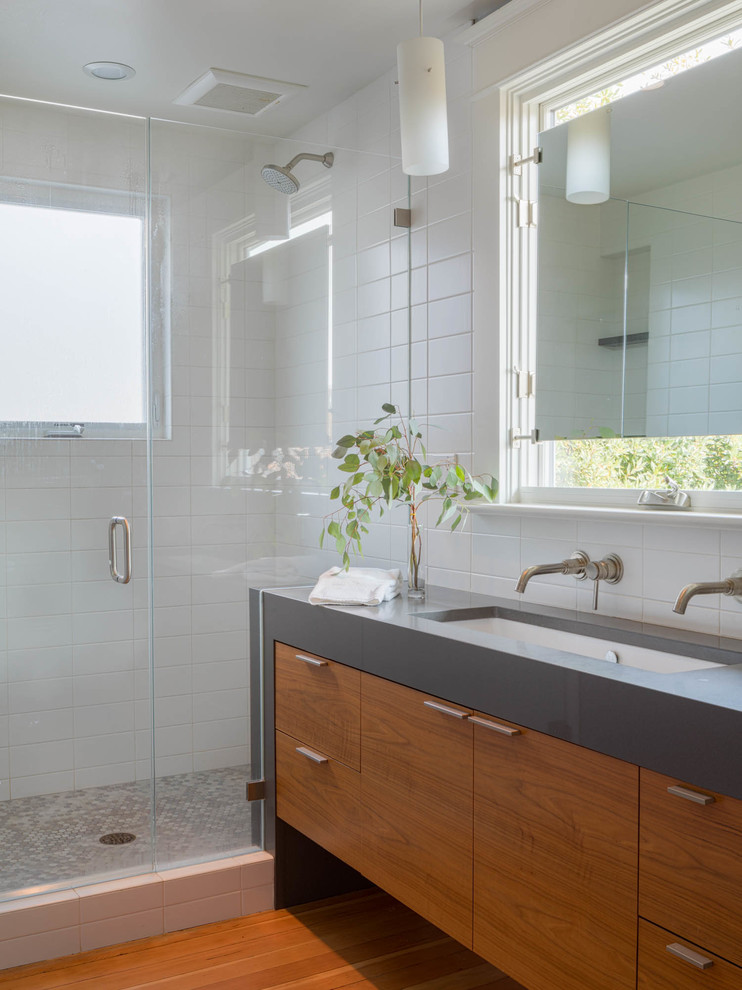 Photo of a contemporary master bathroom in Seattle with flat-panel cabinets, medium wood cabinets, an alcove shower, white tile, white walls, medium hardwood floors, a trough sink, brown floor, a hinged shower door and grey benchtops.