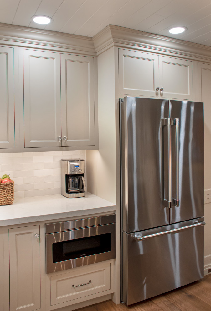 Transitional medium tone wood floor, brown floor and shiplap ceiling kitchen photo with shaker cabinets, beige cabinets, quartz countertops, white backsplash, ceramic backsplash, stainless steel appliances and white countertops