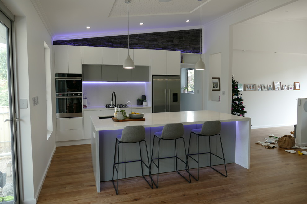 This is an example of a mid-sized modern galley open plan kitchen in Adelaide with an undermount sink, flat-panel cabinets, white cabinets, quartz benchtops, white splashback, stone slab splashback, stainless steel appliances, medium hardwood floors, with island, brown floor and white benchtop.