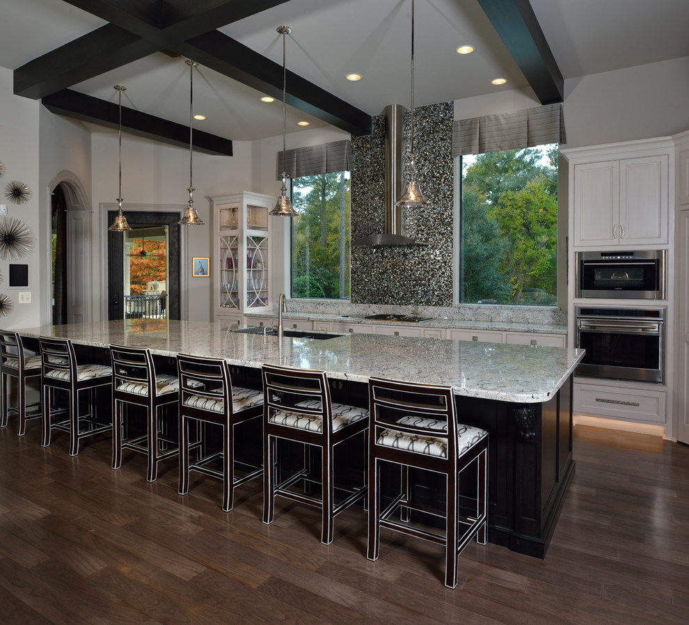Expansive transitional l-shaped open plan kitchen in Houston with recessed-panel cabinets, white cabinets, with island, a drop-in sink, metallic splashback, stainless steel appliances, light hardwood floors, glass tile splashback and granite benchtops.