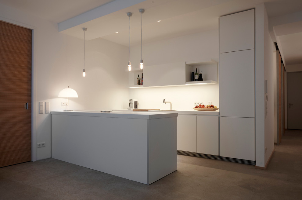 This is an example of a mid-sized contemporary single-wall open plan kitchen in Munich with a drop-in sink, flat-panel cabinets, white cabinets, white splashback, concrete floors and a peninsula.