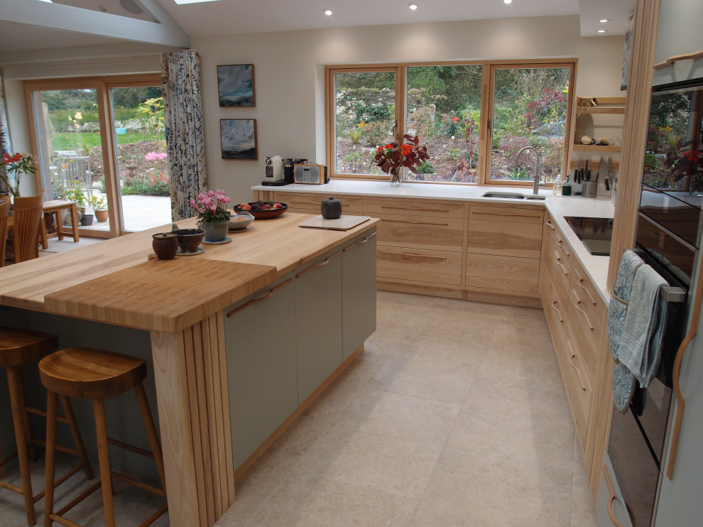 Inspiration for a large contemporary l-shaped open plan kitchen in Cornwall with a submerged sink, flat-panel cabinets, light wood cabinets, engineered stone countertops, blue splashback, black appliances, limestone flooring, an island, beige floors, white worktops, exposed beams and feature lighting.