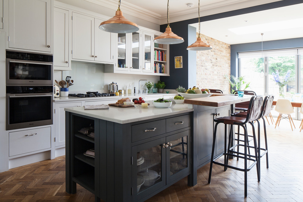 Design ideas for a classic kitchen/diner in Sussex with shaker cabinets, white cabinets, white splashback, glass sheet splashback, stainless steel appliances, dark hardwood flooring, an island, brown floors and white worktops.