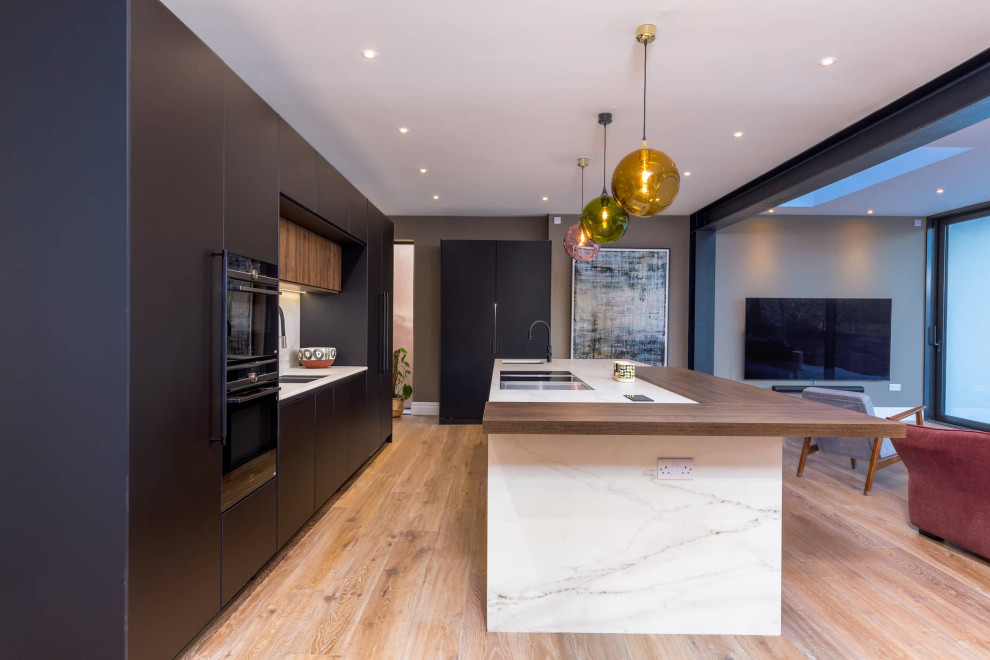 Large modern single-wall open plan kitchen in Other with flat-panel cabinets, black cabinets, quartzite benchtops, white splashback, black appliances, light hardwood floors, with island, brown floor and white benchtop.