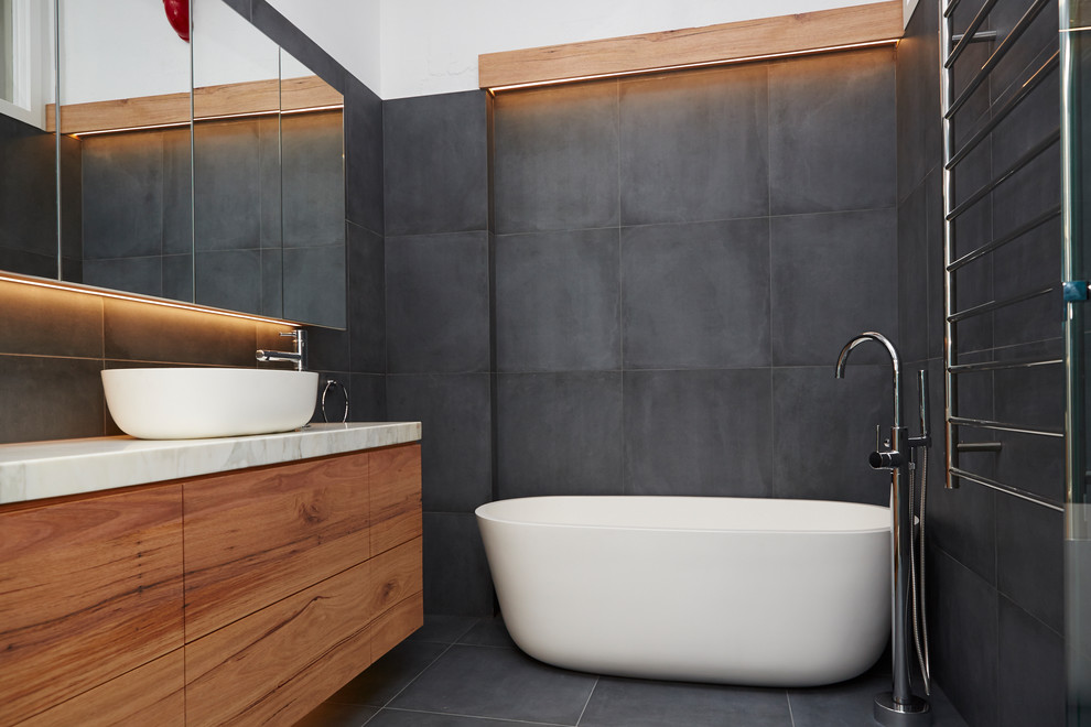 Photo of a mid-sized modern master bathroom in Melbourne with recessed-panel cabinets, light wood cabinets, a freestanding tub, an open shower, a wall-mount toilet, black tile, porcelain tile, black walls, porcelain floors, a wall-mount sink, marble benchtops, black floor, a hinged shower door and white benchtops.