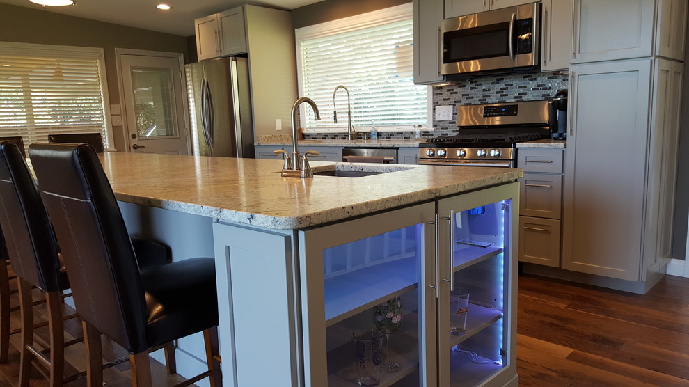 Photo of a mid-sized transitional galley open plan kitchen in Indianapolis with an undermount sink, shaker cabinets, grey cabinets, granite benchtops, grey splashback, glass tile splashback, stainless steel appliances, medium hardwood floors and with island.