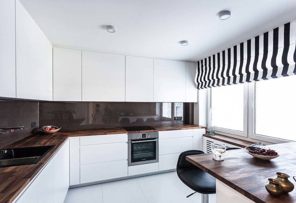 Inspiration for a mid-sized contemporary l-shaped separate kitchen in Berlin with a drop-in sink, flat-panel cabinets, white cabinets, wood benchtops, black splashback, glass sheet splashback, stainless steel appliances, a peninsula, white floor and brown benchtop.