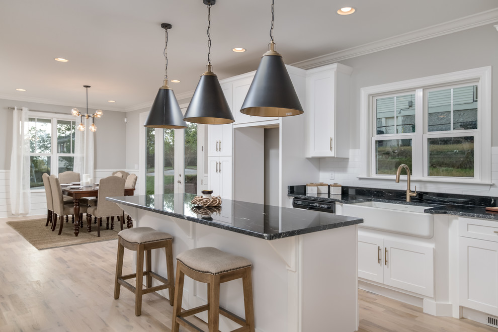 Inspiration for a mid-sized arts and crafts l-shaped open plan kitchen in Other with a farmhouse sink, shaker cabinets, white cabinets, granite benchtops, white splashback, ceramic splashback, stainless steel appliances, light hardwood floors, with island, beige floor and black benchtop.
