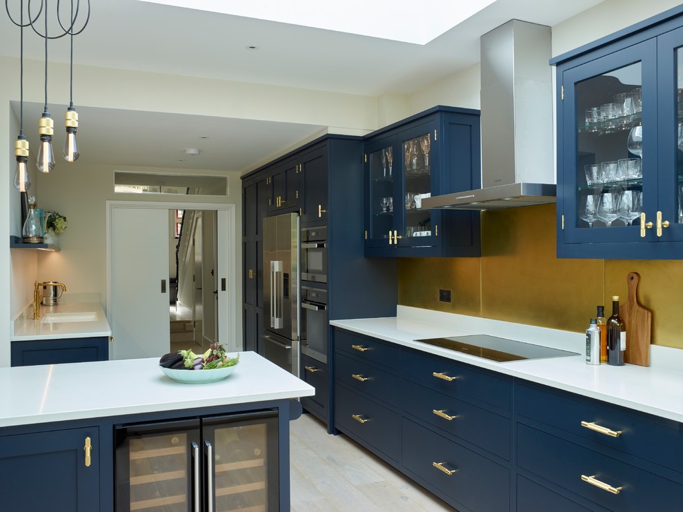This is an example of a mid-sized contemporary galley eat-in kitchen in London with a double-bowl sink, shaker cabinets, blue cabinets, solid surface benchtops, stainless steel appliances, light hardwood floors, with island and white benchtop.