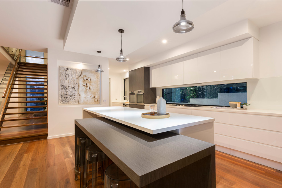 Photo of a contemporary kitchen in Melbourne with flat-panel cabinets, white cabinets, window splashback, stainless steel appliances, medium hardwood floors, multiple islands and white benchtop.