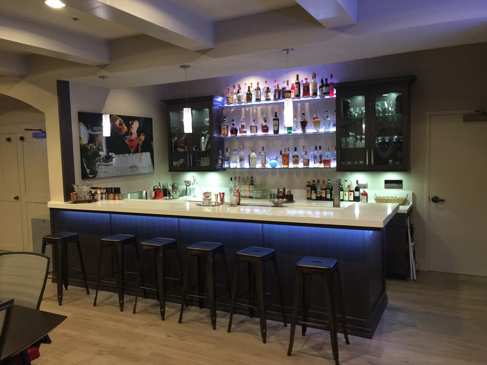 This is an example of a large modern u-shaped seated home bar in Miami with a drop-in sink, glass-front cabinets, dark wood cabinets, marble benchtops, light hardwood floors, brown floor and white benchtop.