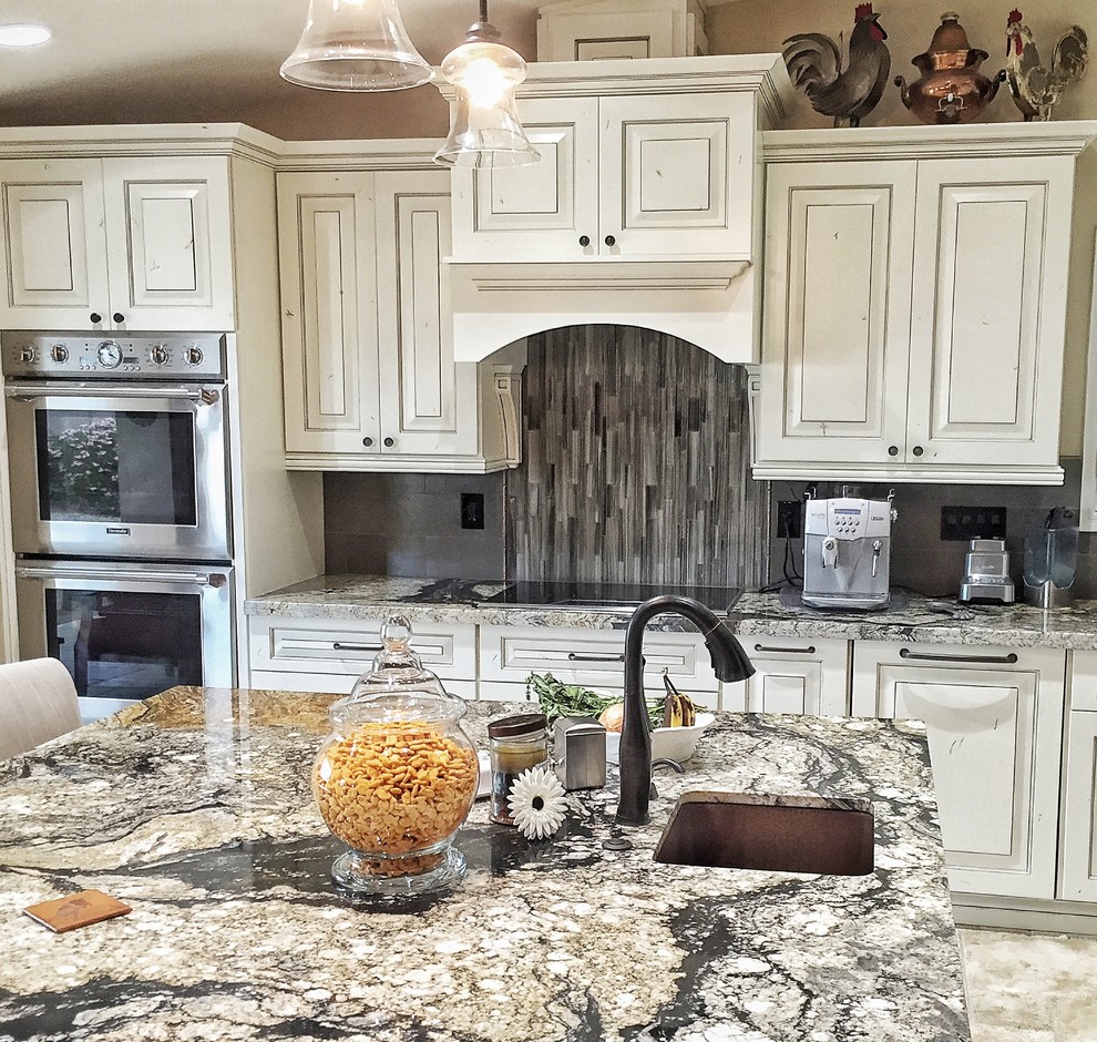 Photo of a large traditional l-shaped eat-in kitchen in Phoenix with a farmhouse sink, raised-panel cabinets, white cabinets, granite benchtops, grey splashback, glass sheet splashback, stainless steel appliances, travertine floors, with island, beige floor and multi-coloured benchtop.