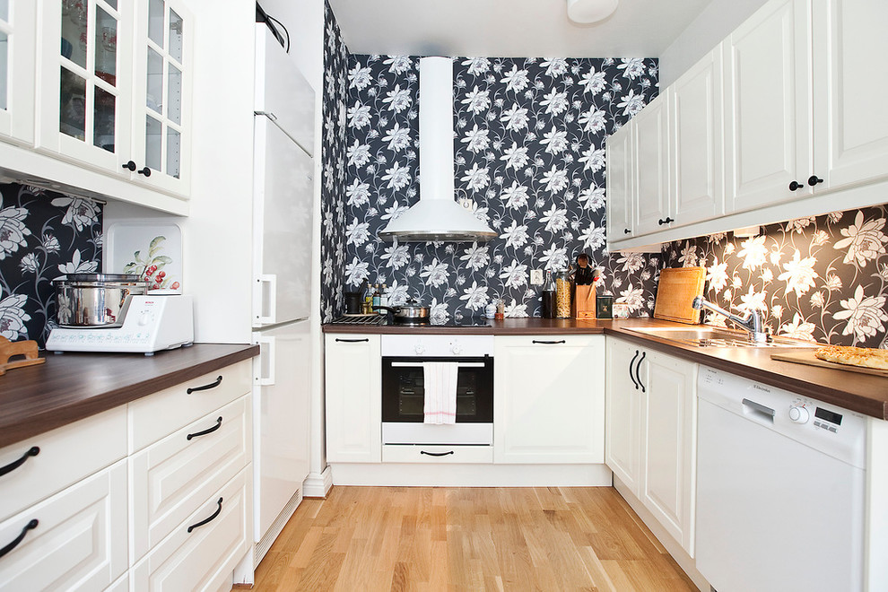 This is an example of a transitional u-shaped separate kitchen in Malmo with a drop-in sink, raised-panel cabinets, white cabinets, wood benchtops, white appliances, light hardwood floors and no island.