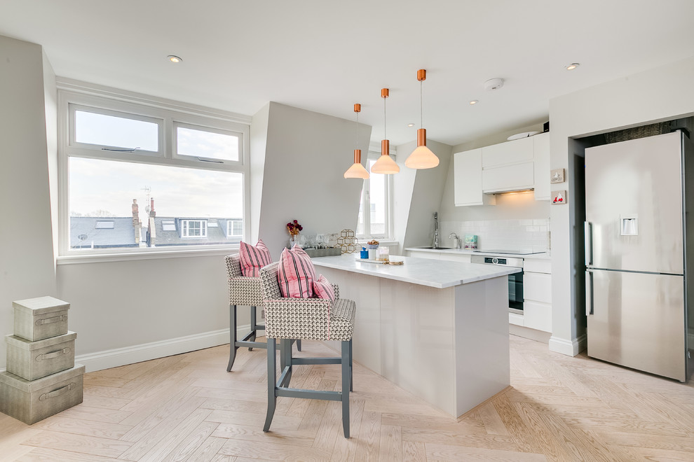 This is an example of a small contemporary galley open plan kitchen in London with a single-bowl sink, flat-panel cabinets, white cabinets, solid surface benchtops, white splashback, black appliances, light hardwood floors, subway tile splashback, a peninsula, beige floor and white benchtop.