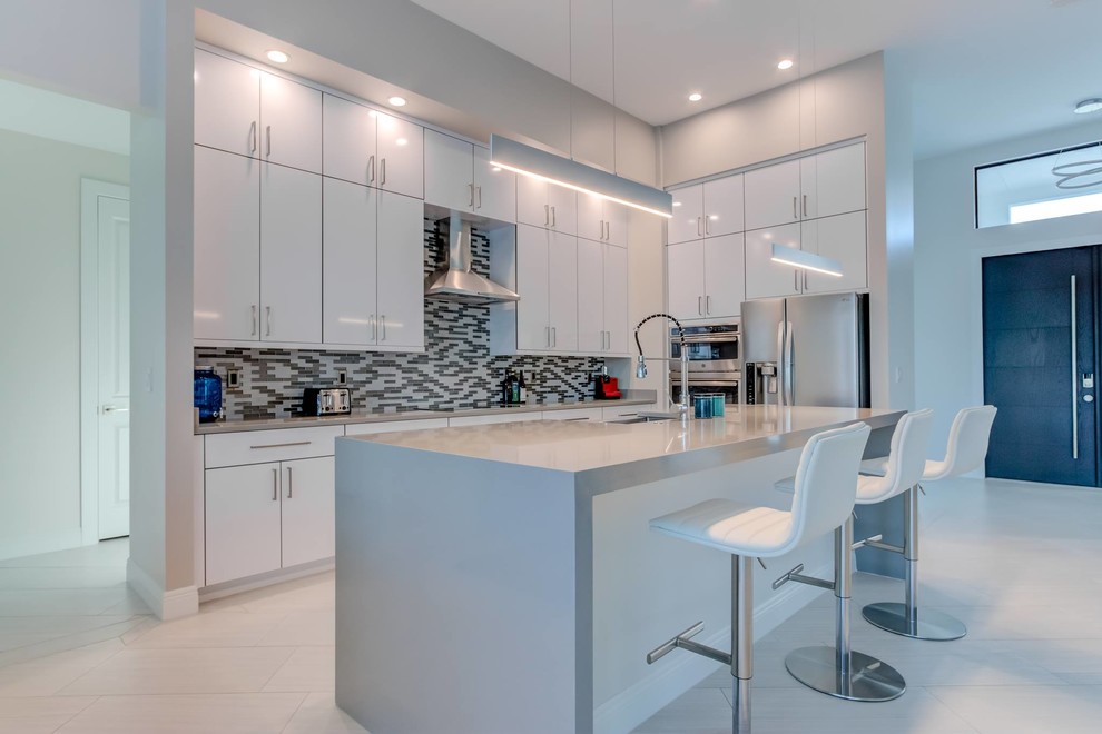 This is an example of a large modern single-wall open plan kitchen in Other with a farmhouse sink, flat-panel cabinets, white cabinets, quartz benchtops, multi-coloured splashback, glass tile splashback, stainless steel appliances, with island, white floor and grey benchtop.