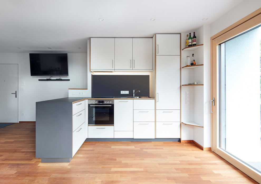 Offene, Mittelgroße Moderne Küche in L-Form mit Waschbecken, flächenbündigen Schrankfronten, weißen Schränken, Arbeitsplatte aus Holz, Küchenrückwand in Grau, schwarzen Elektrogeräten, hellem Holzboden, Halbinsel, braunem Boden und grauer Arbeitsplatte in Sonstige