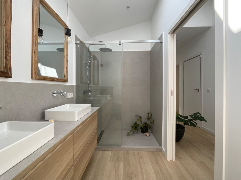 Mid-sized modern master bathroom in Barcelona with white cabinets, a curbless shower, a wall-mount toilet, gray tile, white walls, laminate floors, a vessel sink, tile benchtops, brown floor, a sliding shower screen, grey benchtops, a double vanity and a floating vanity.